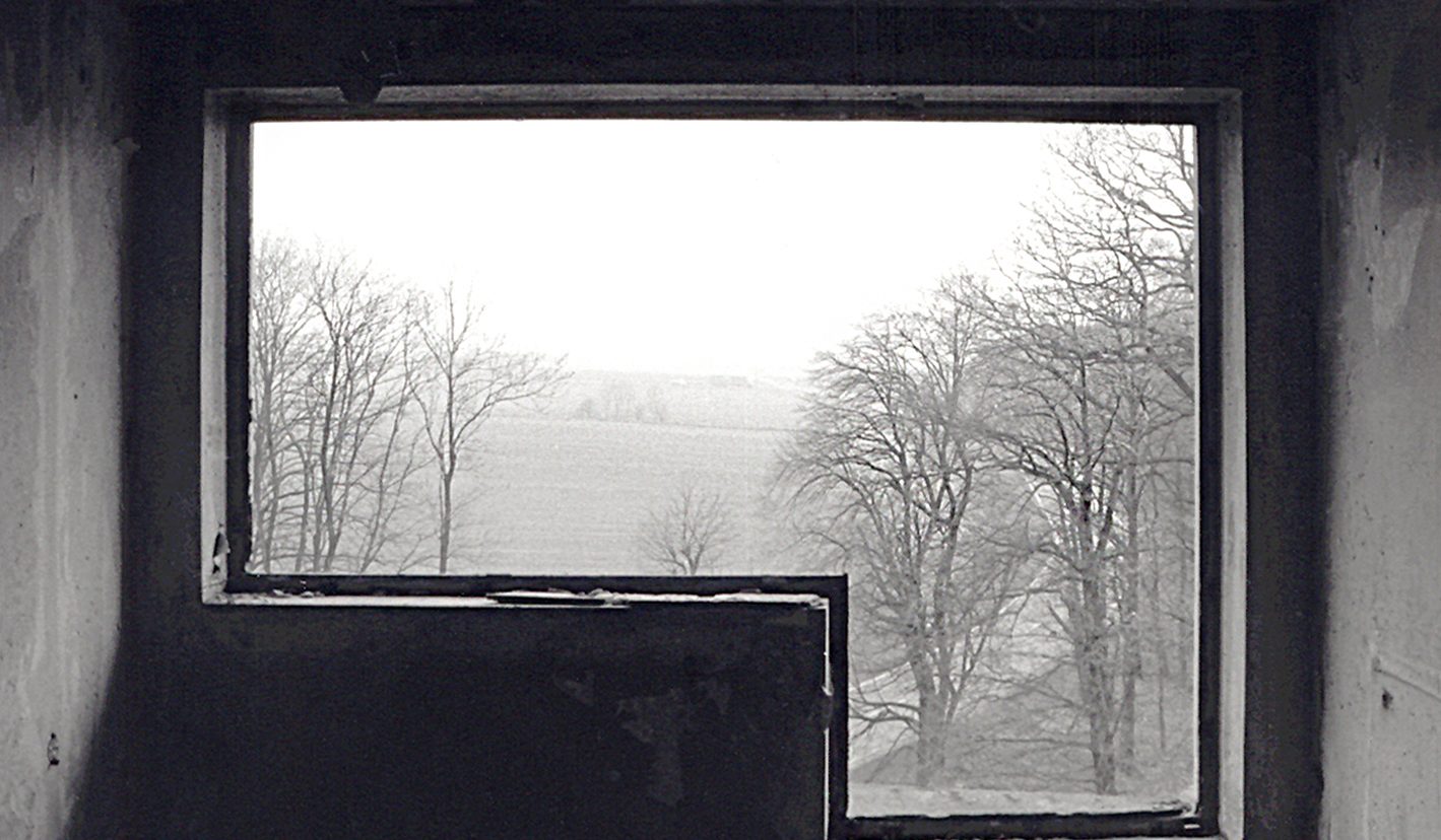Schwarzweiß Fotografie von einem leerstehenden Raum, nach außen durch die Fenster-Öffnung, in der kein Glas mehr vorhanden ist. Der Blick fällt auf ein Feld und Bäume ohne Laub im Herbst.