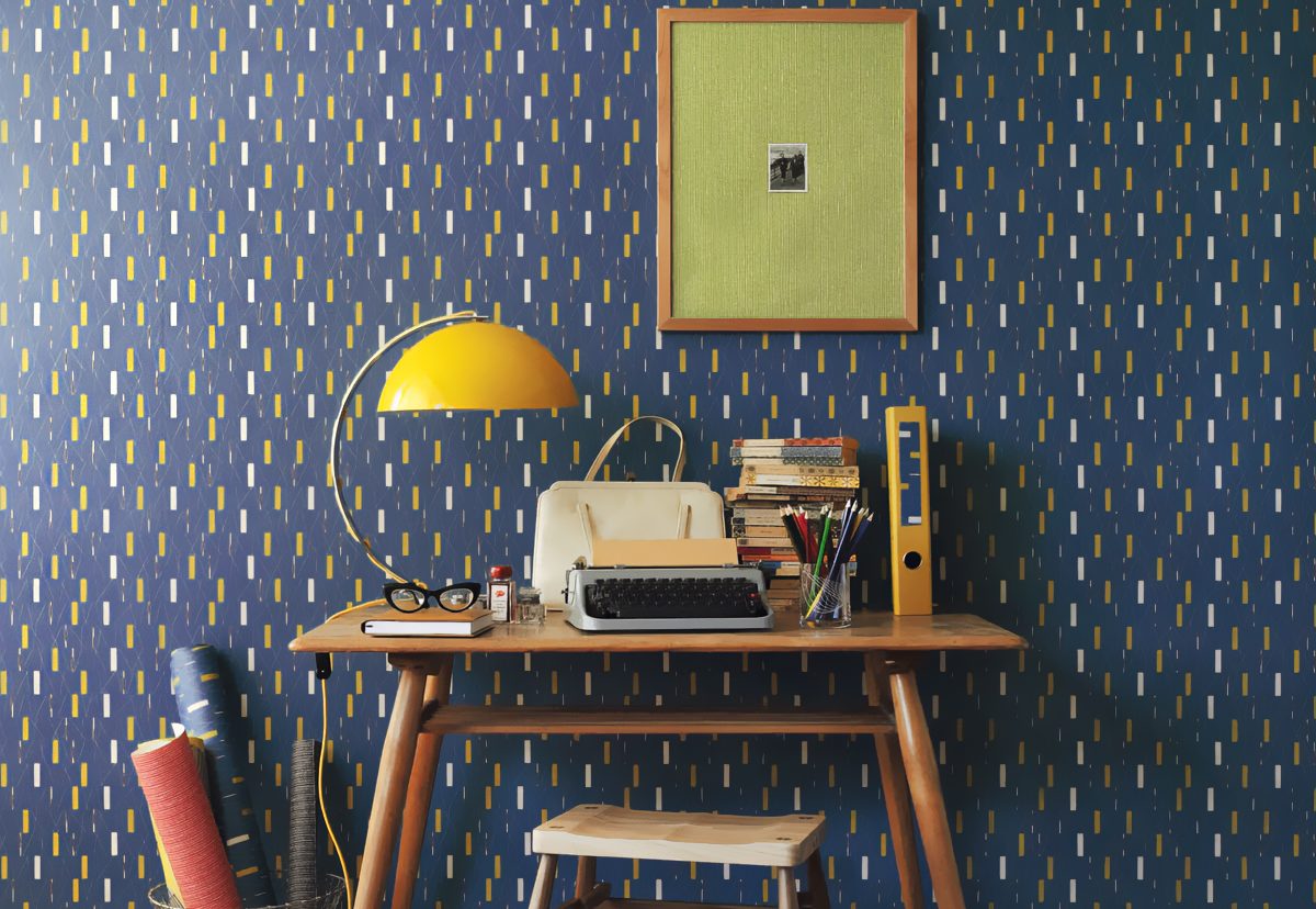Ein alter Retro Vintage Schreibtisch vor einer Wand mit blauer Muster Tapete im Stil der 60iger Jahre. Auf dem Schreibtisch liegen verschiedene Skizzenbücher und Schreibutensilien und am Rand steht eine gelbe Kugellampe und eine Schreibmaschine.